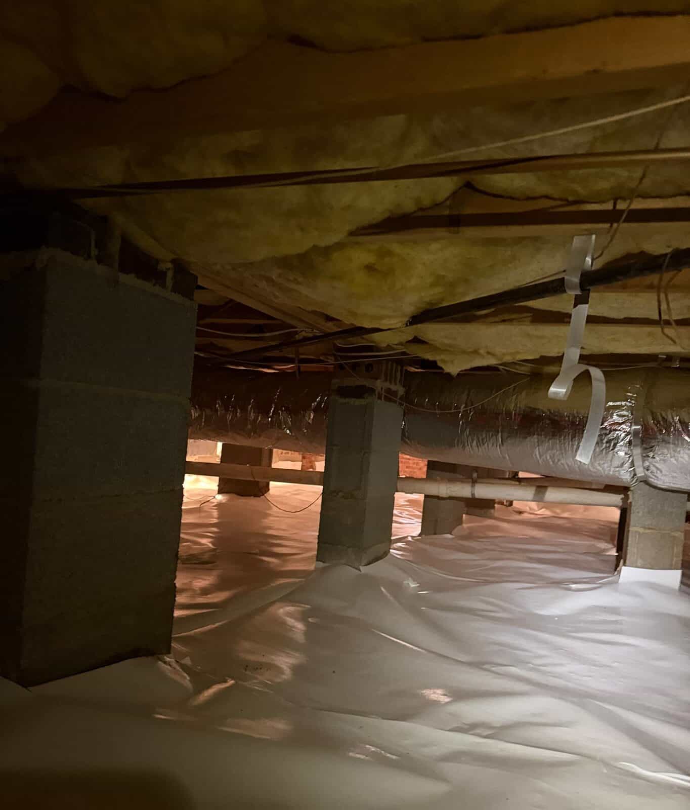 Block piers stand on a crawl space floor covered with white vapor barrier under insulated joists.