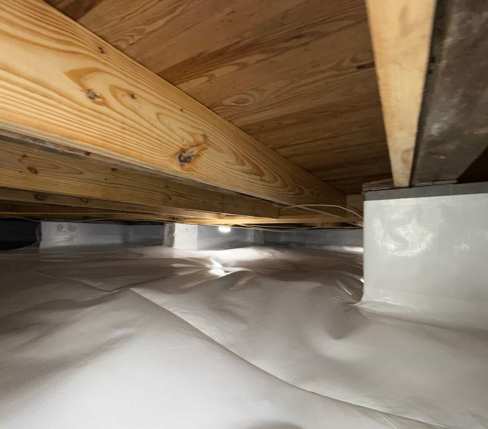 Crawl space with white liner on the floor, a wrapped pier and new lumber joists above.