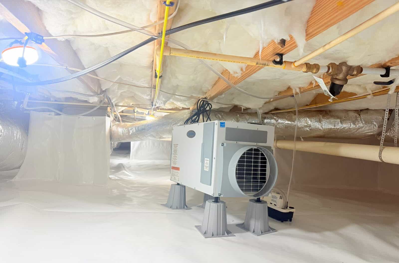 Dehumidifier on stands in a crawl space lined with white liner, with ducts and insulation overhead.