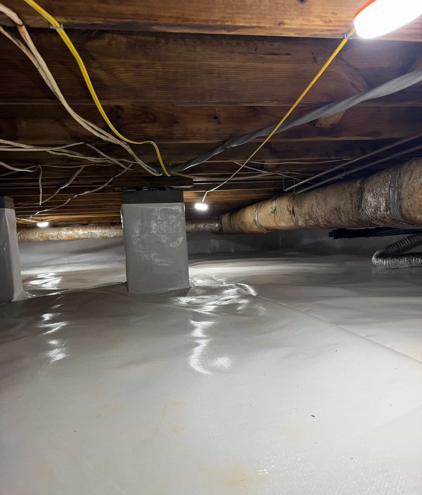 Lined crawl space with wrapped piers and a work light under the floor joists.