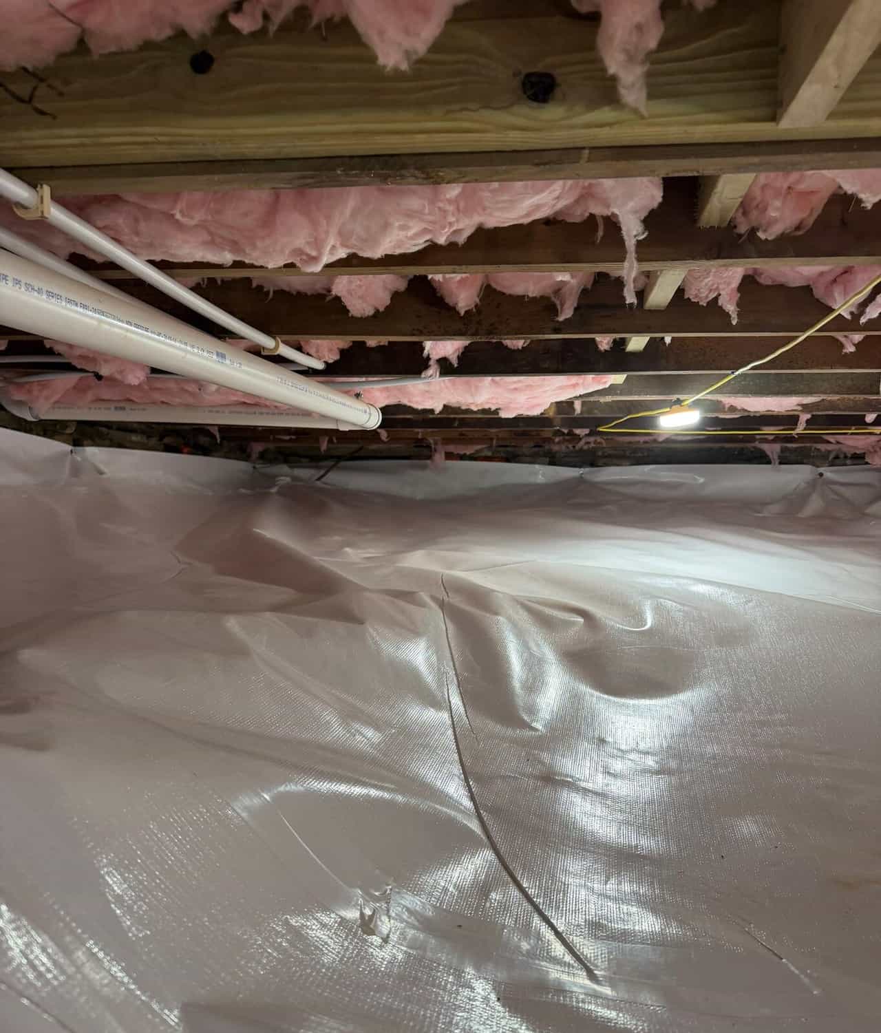 Crawl space floor covered in white liner with a taped seam below plumbing pipes and insulated joists.