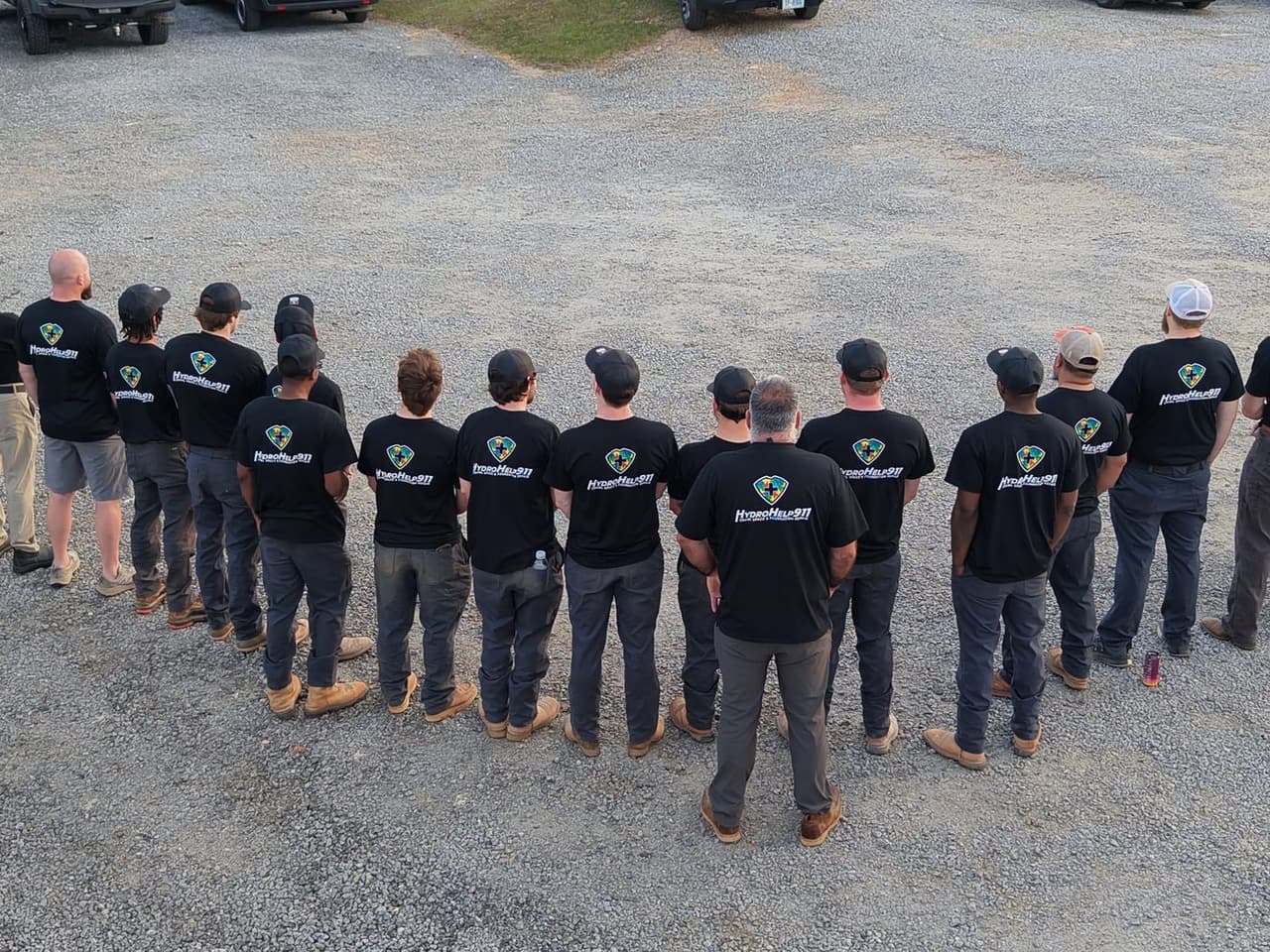 The HydroHelp911 team standing in a row on a gravel lot, backs to the camera, in matching black HydroHelp911 shirts