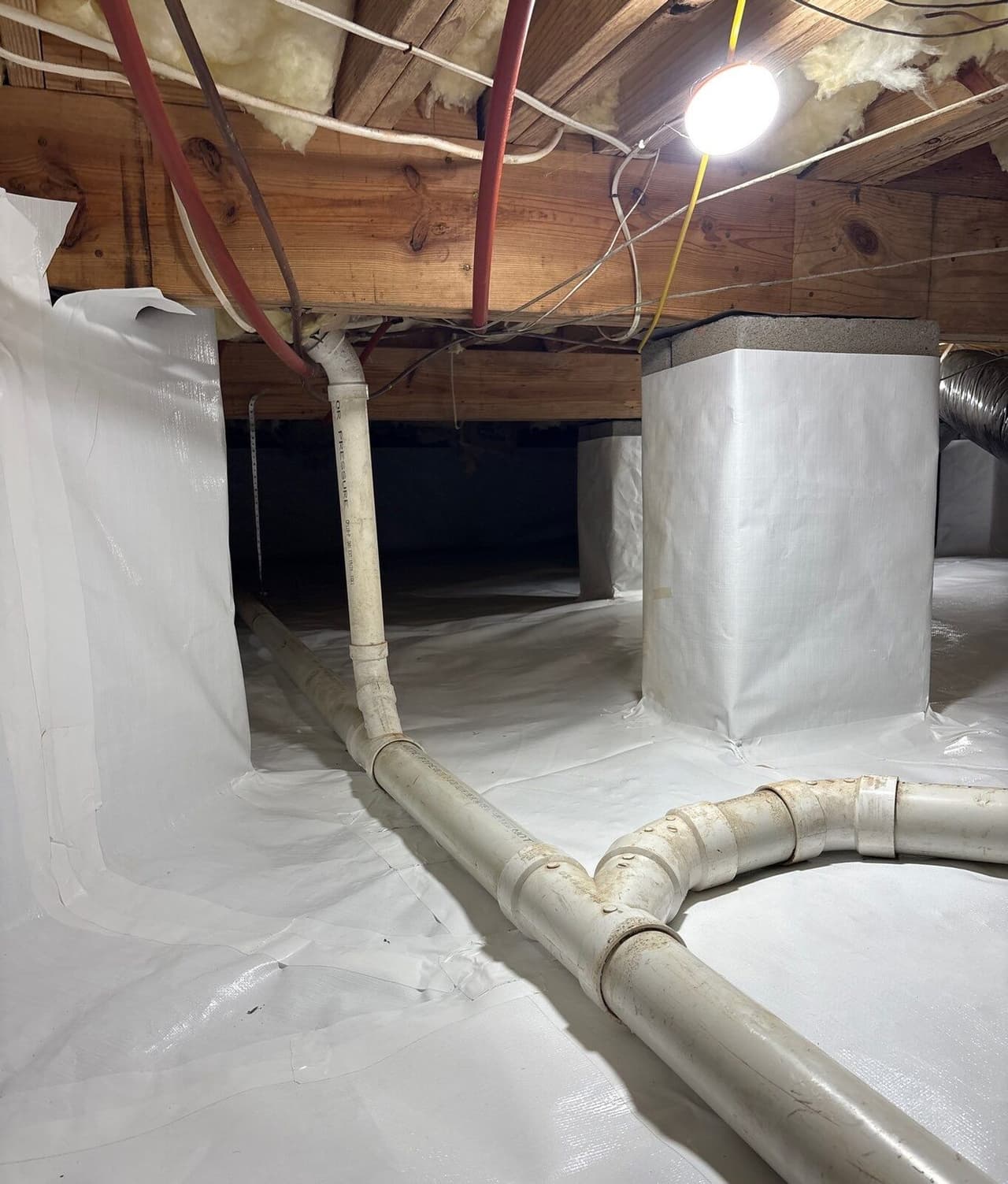 Crawl space with white liner on walls and floor, wrapped piers and white drain pipes.
