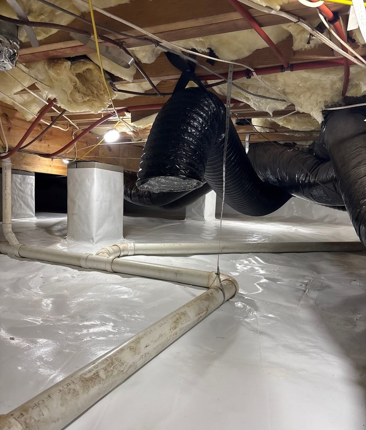 Lined crawl space with wrapped piers, white drain pipes and black ducts under the floor joists.