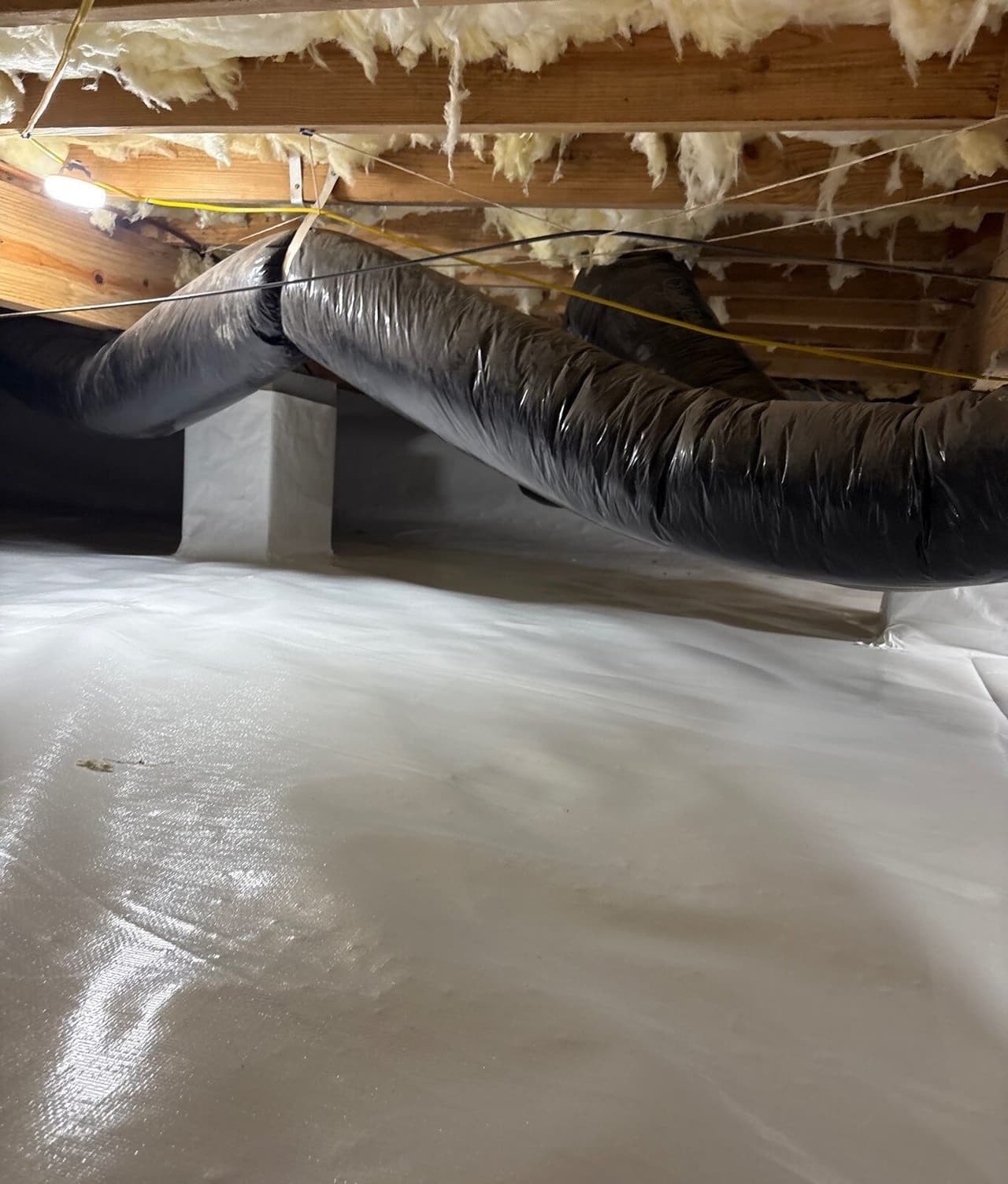 Crawl space floor covered in white liner with a wrapped pier and ducts overhead.
