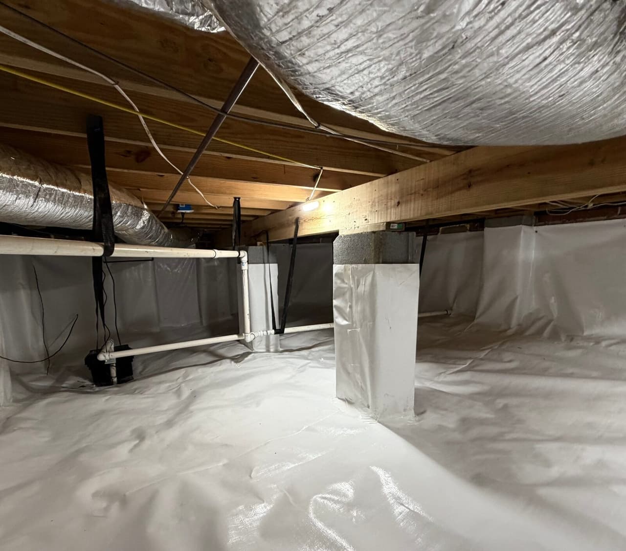 Crawl space with white liner on floor and walls, a wrapped pier and a small pump near a pipe.