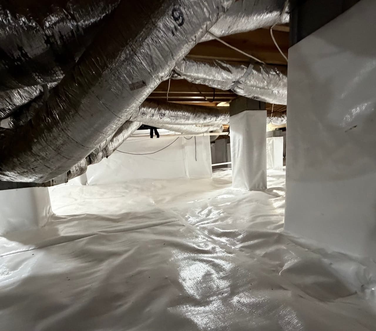 Low crawl space lined in white plastic with wrapped piers beneath insulated ducts.
