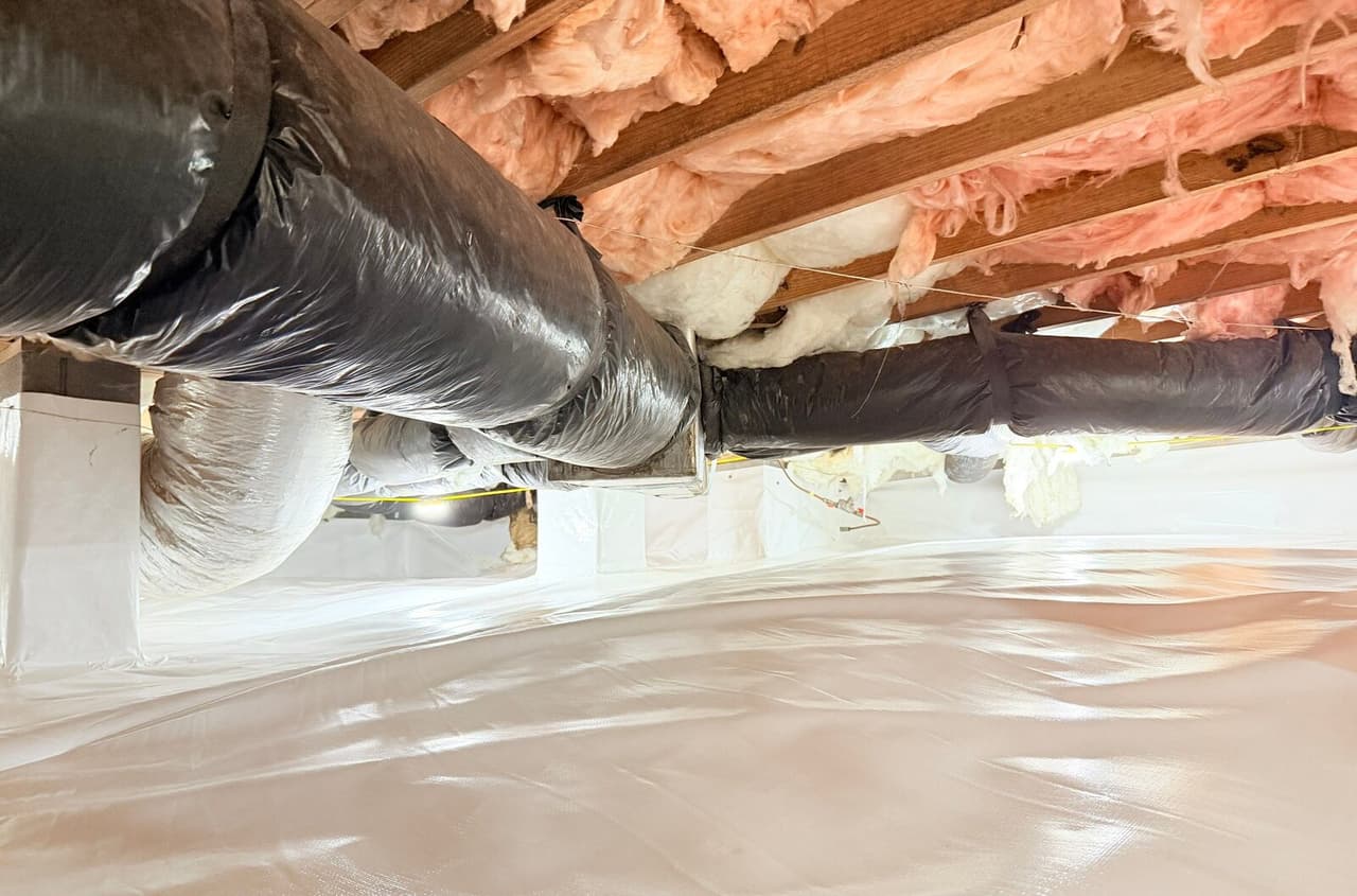Crawl space floor covered in white liner with wrapped piers under ducts and insulation.