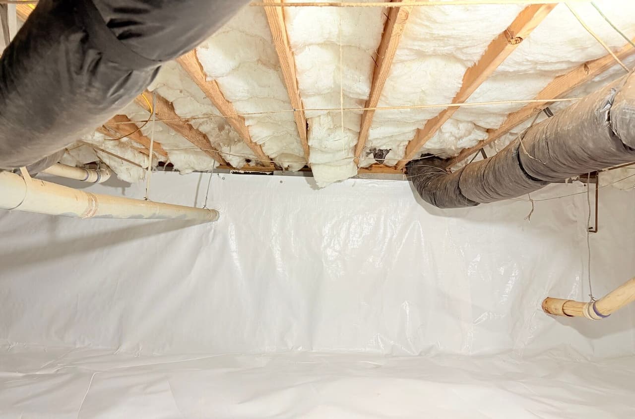 White liner lining a crawl space wall and floor beneath insulated joists and ducts.