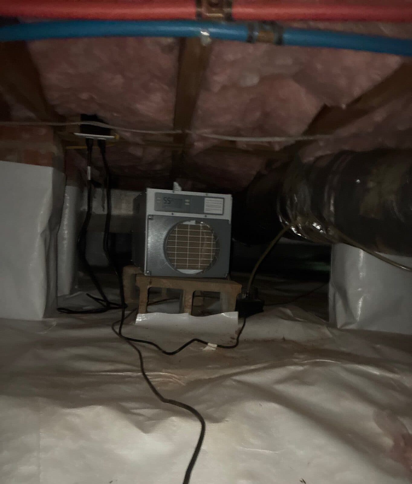 Dehumidifier on blocks in a lined crawl space with wrapped piers and a duct nearby.