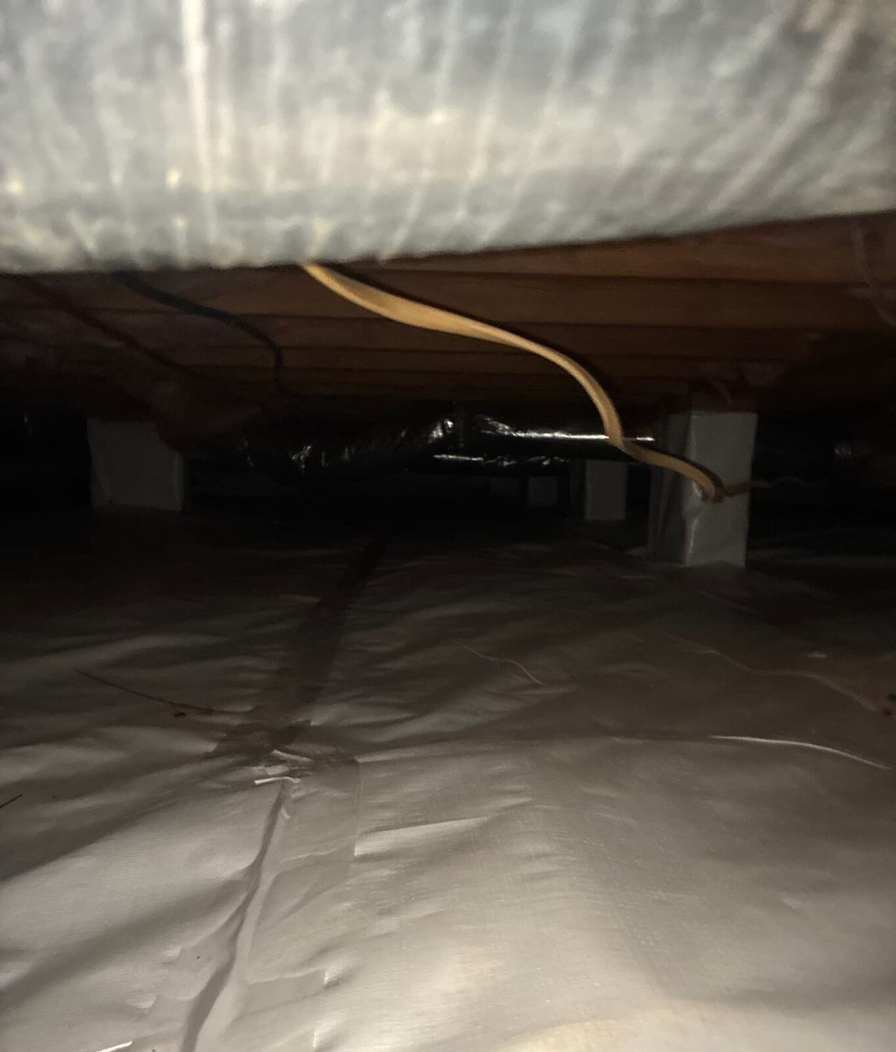 Dim crawl space with taped white liner across the floor and wrapped piers in the distance.