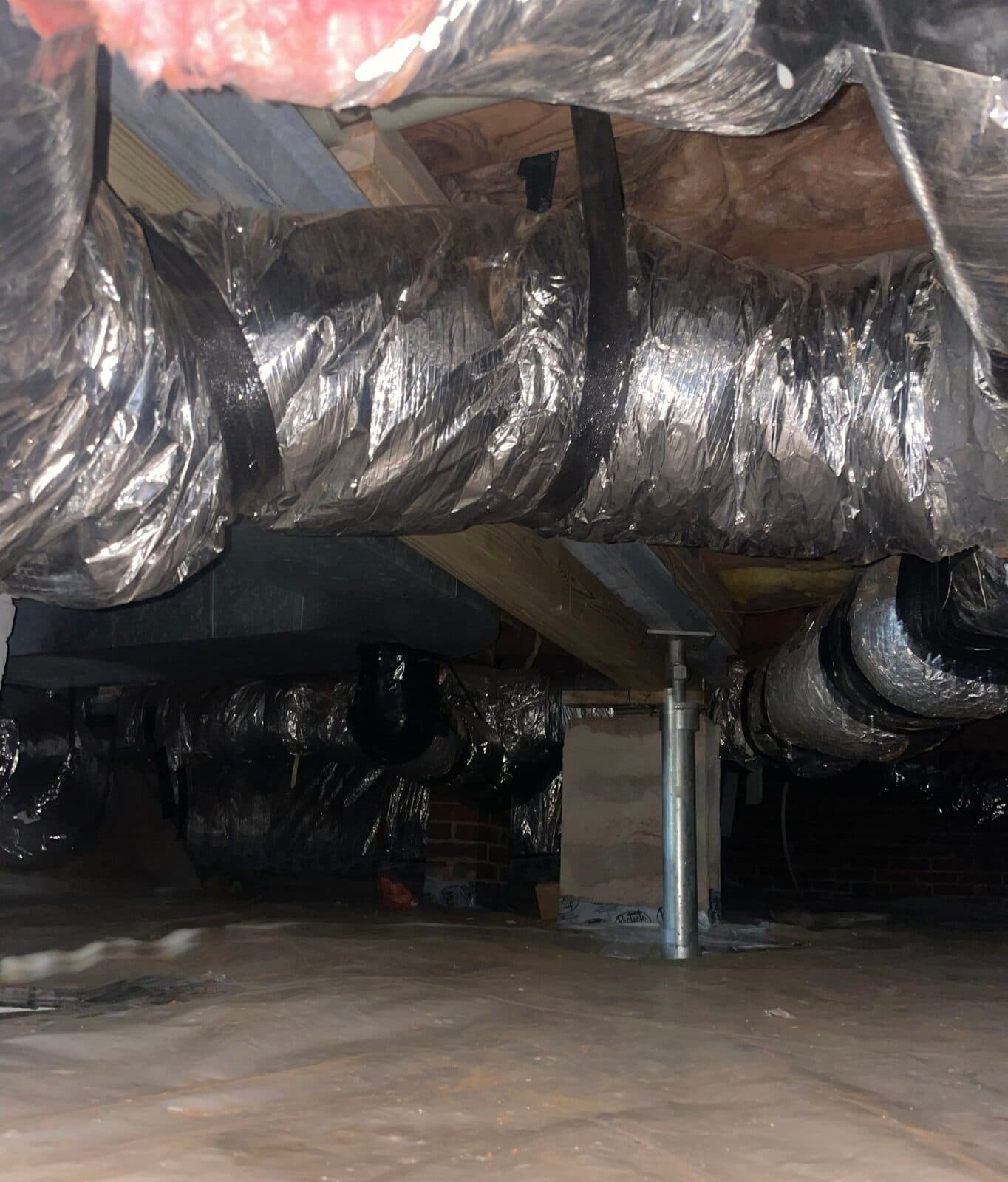 Crawl space with a steel support post under a wood beam, foil ducts overhead and plastic covering the floor.