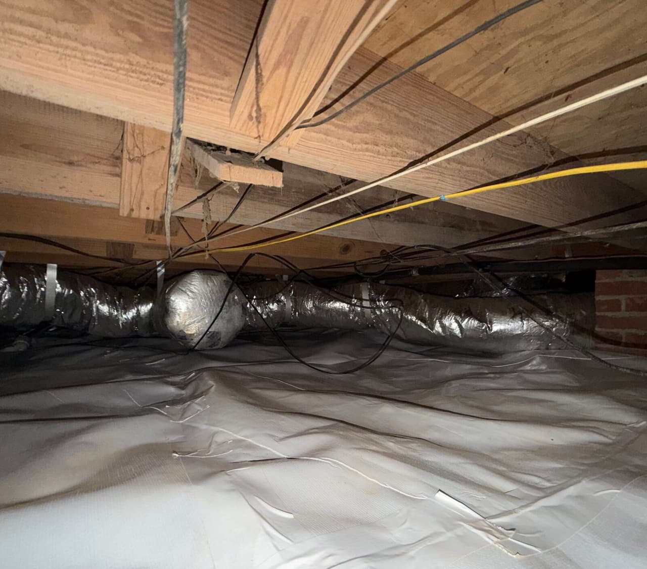 Crawl space floor covered with white liner under wood joists, wiring and insulated ducts.