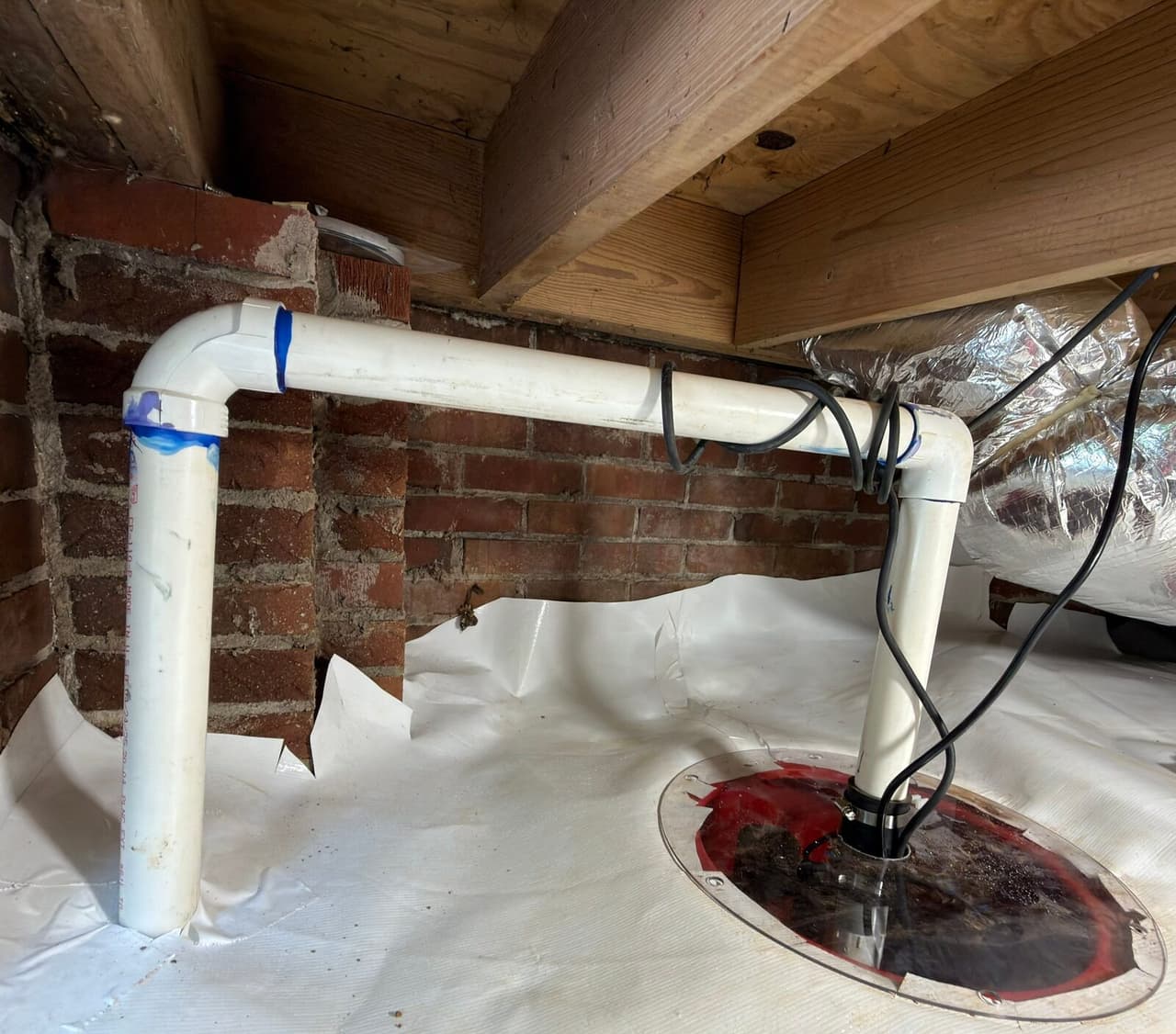 Sump basin with clear lid set in the crawl space liner, with white PVC discharge pipe running along a brick wall.