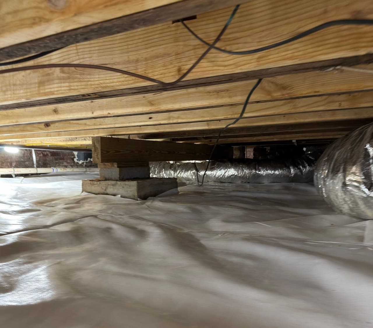 Crawl space floor covered with white vapor barrier liner beneath wood floor joists and a wood support block.