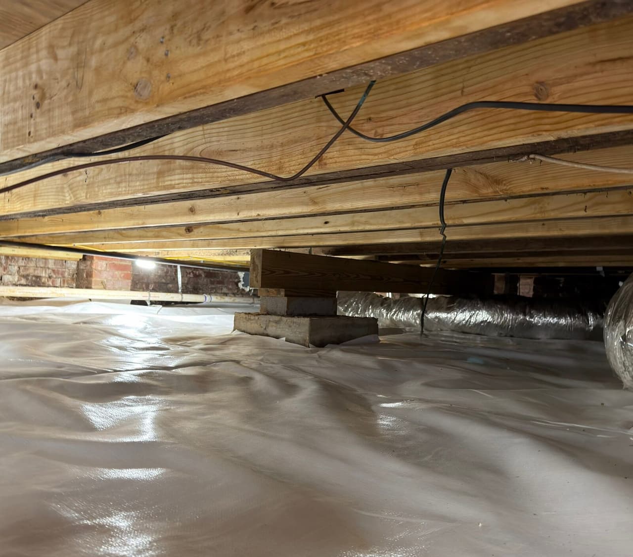 White liner covering a crawl space floor under wood joists, with insulated ducts and brick walls behind.