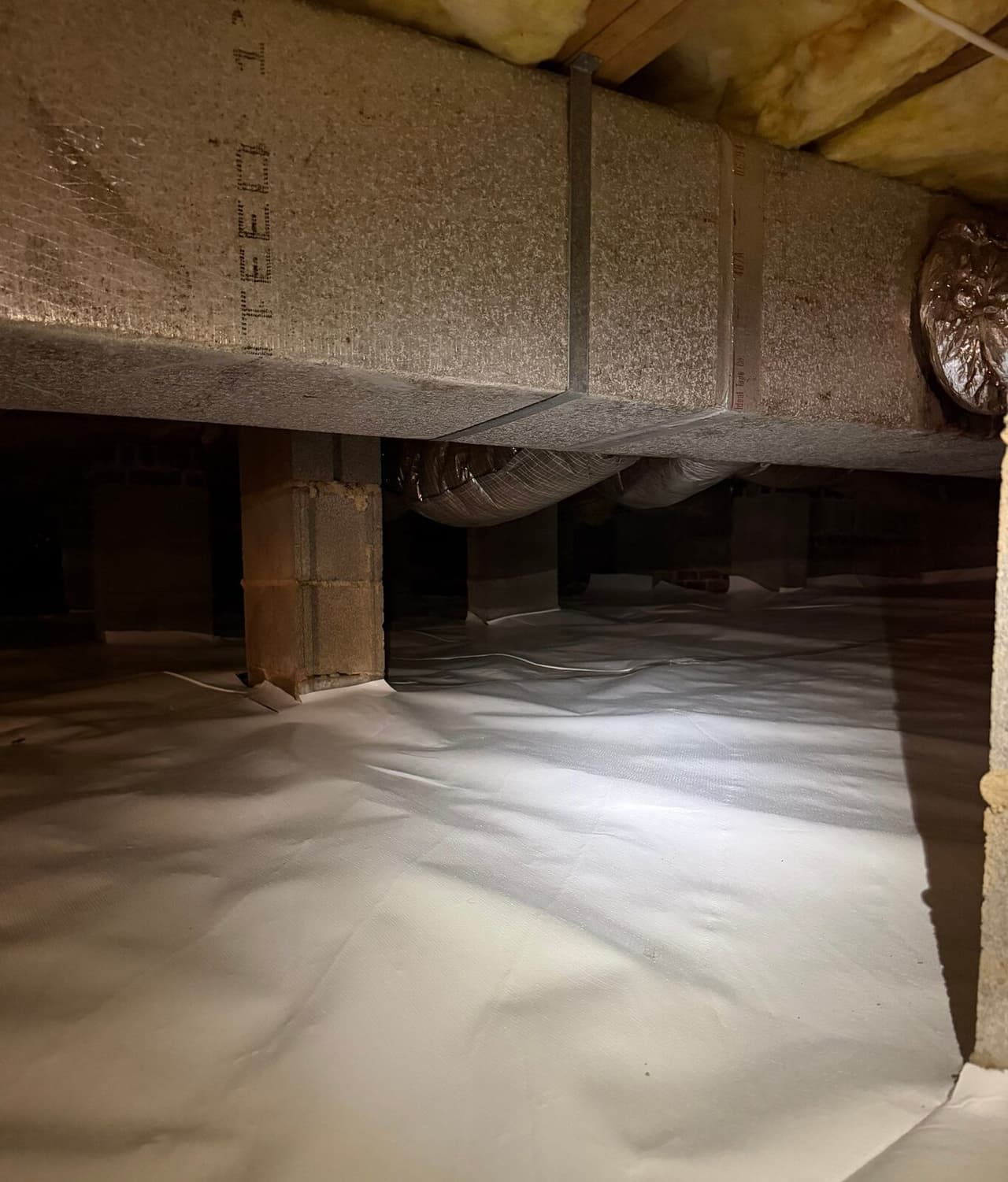 Crawl space with white vapor barrier covering the floor around block piers beneath an insulated duct.