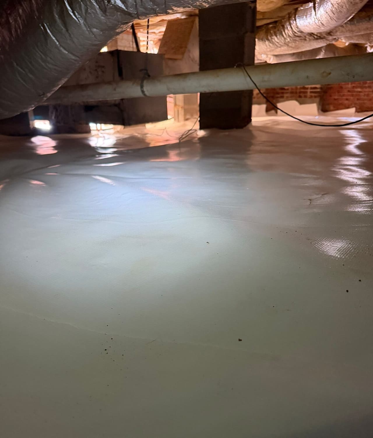 Glossy white vapor barrier covers the crawl space floor below a pipe and duct.