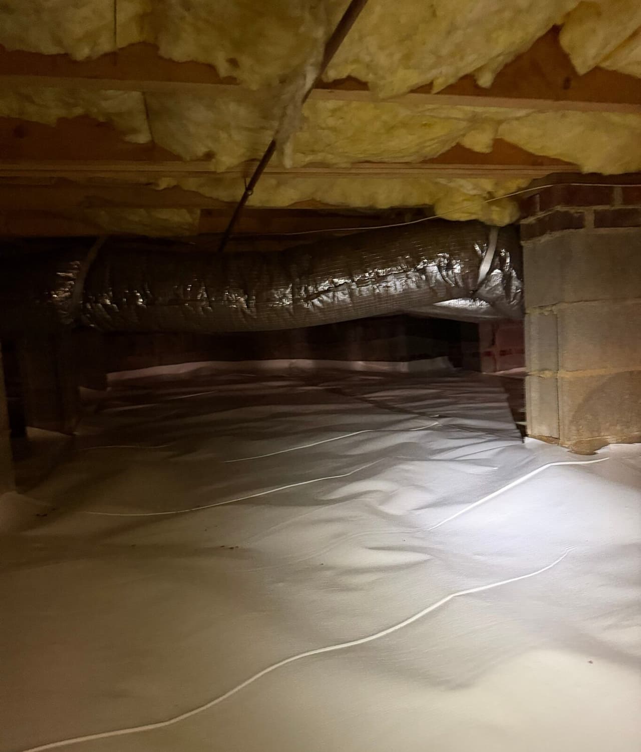 Crawl space with white vapor barrier on the floor beneath insulation and a flexible duct.