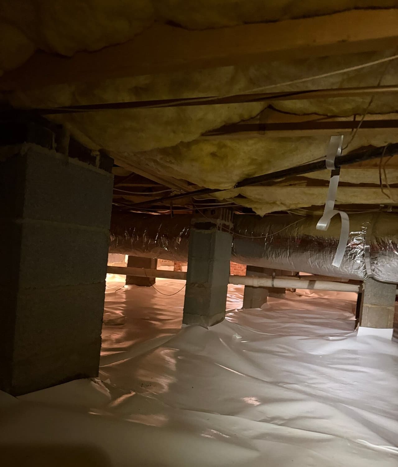 Block piers stand on a crawl space floor covered with white vapor barrier under insulated joists.