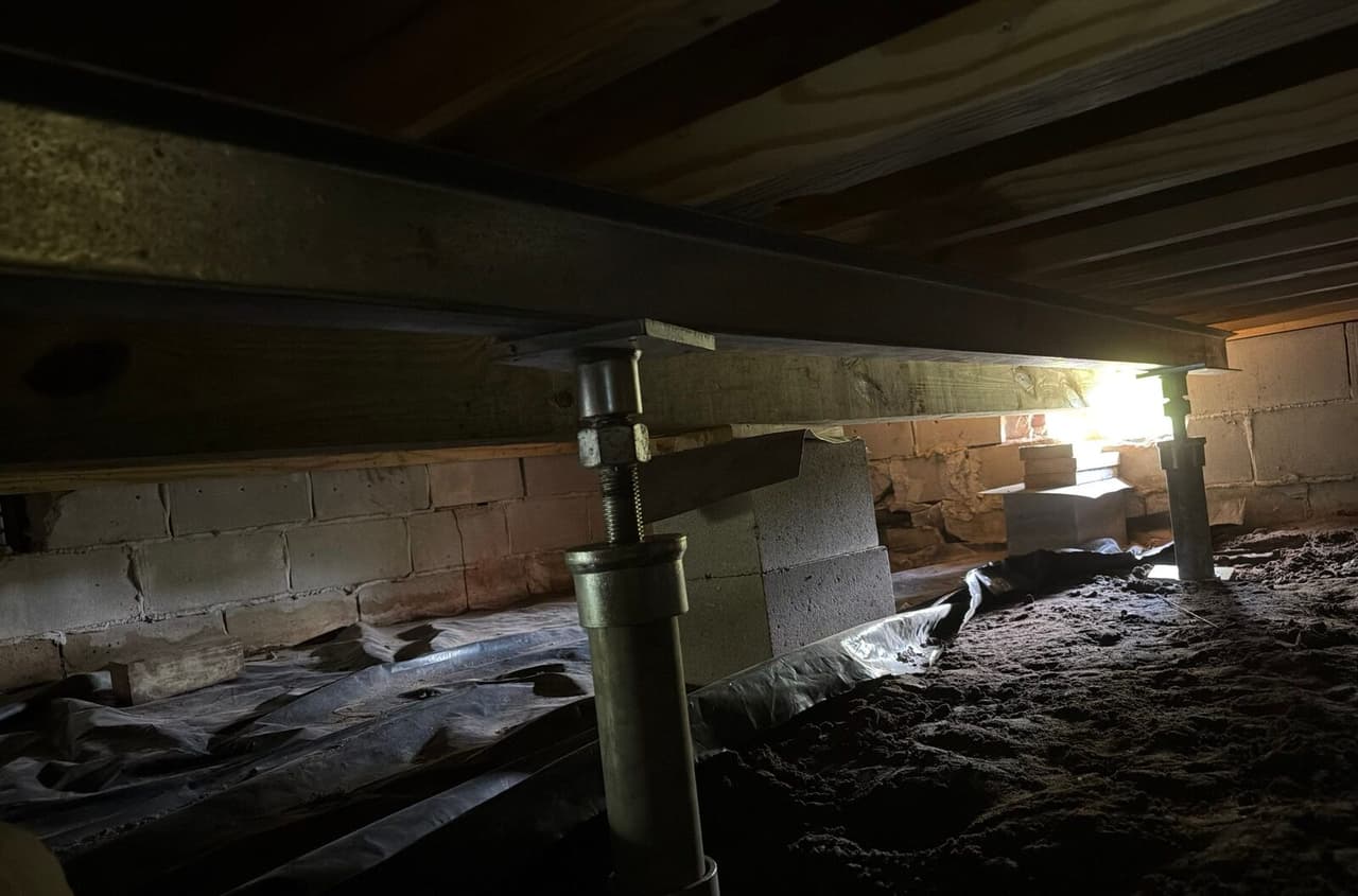 Dim crawl space with two adjustable steel posts supporting a beam near block piers and foundation walls.