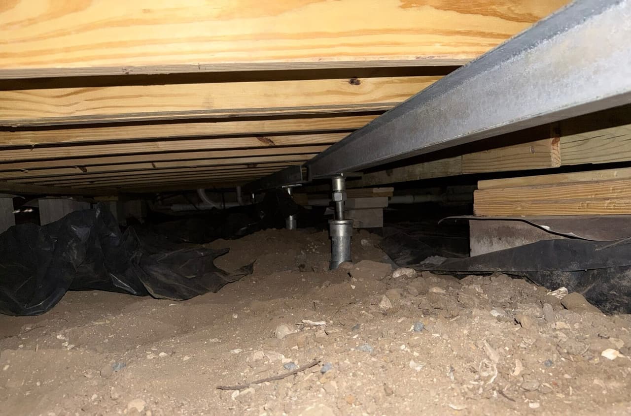 Adjustable steel post holding up a steel beam under wood floor joists in a dirt crawl space.