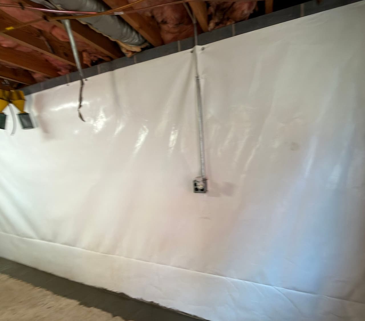 Basement wall covered in white liner below exposed joists, with electrical conduit running down to a box.