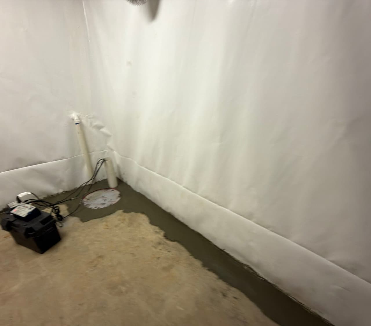 Basement corner with white wall liner, covered sump basin, discharge pipe and a battery backup unit on the floor.