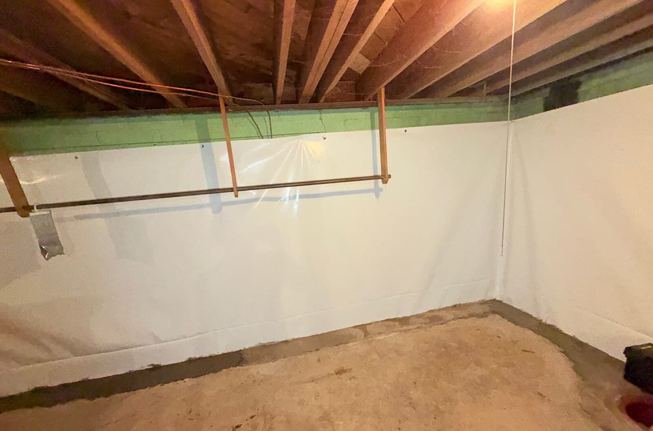 Basement corner with white wall liner fastened to block walls and fresh concrete patch along the floor edge.