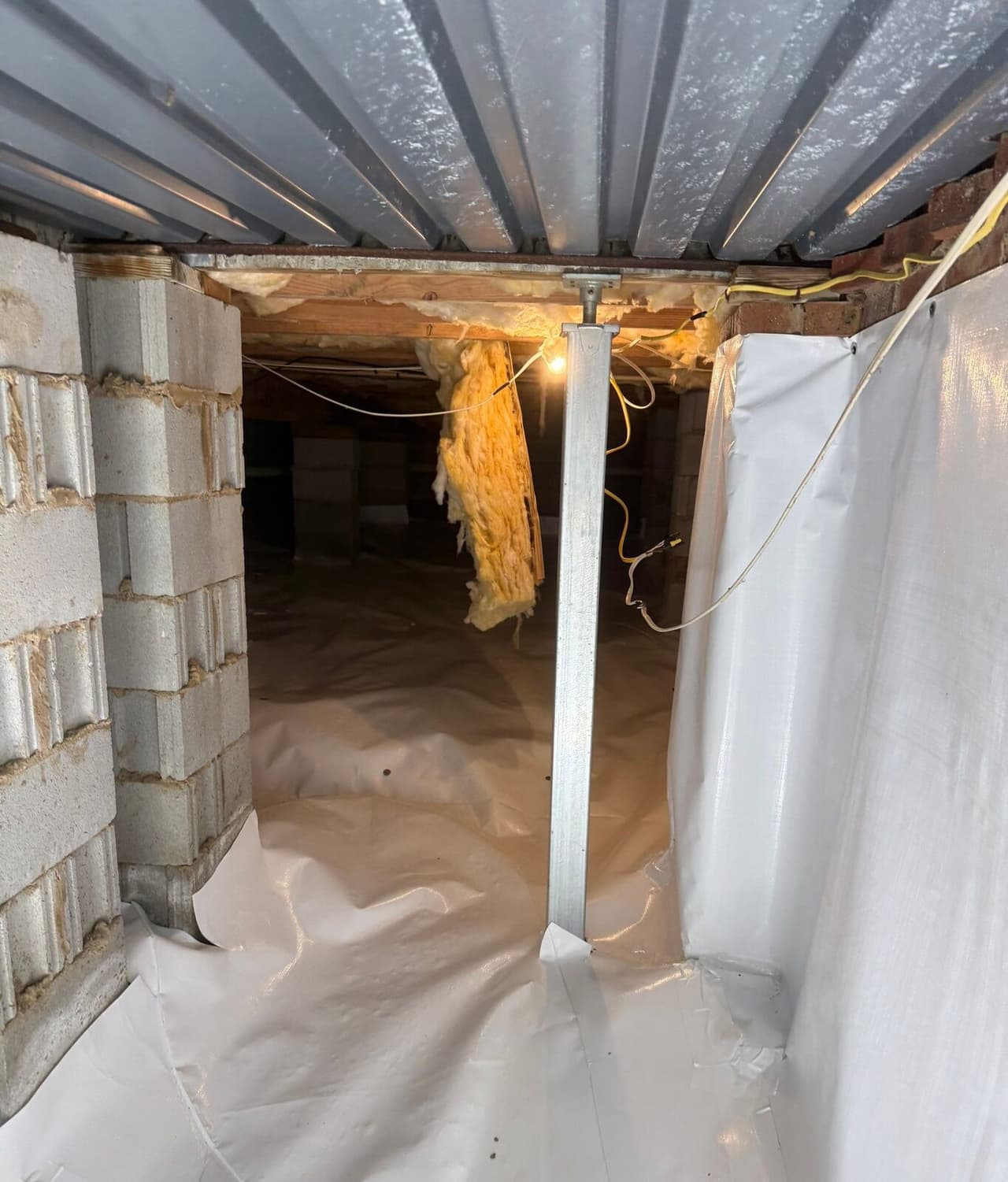 Steel support post between a block pier and a lined wall in a crawl space with white floor liner.