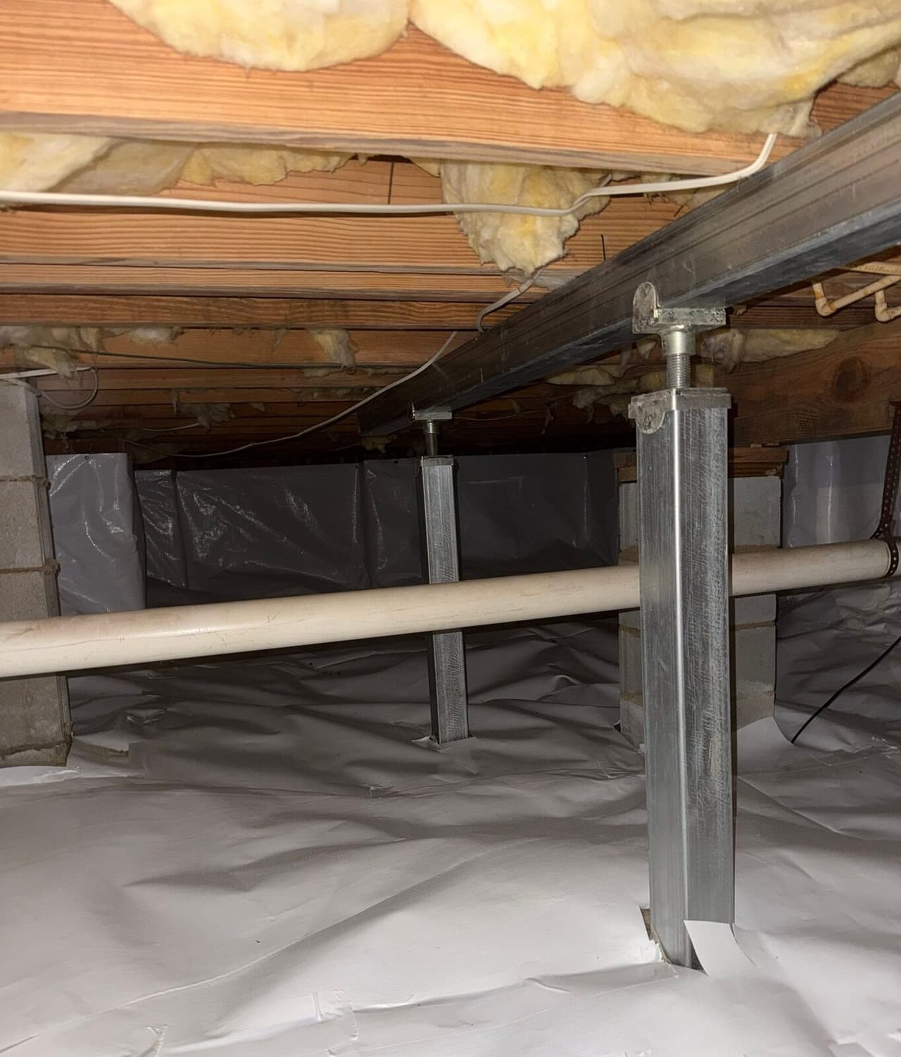 Two steel support posts under a metal beam in a crawl space with white liner on the floor and walls.