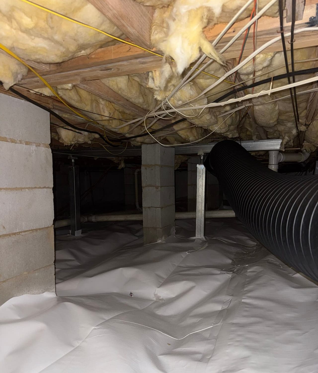 Crawl space with white floor liner, steel support posts, block piers, and a black flexible duct.