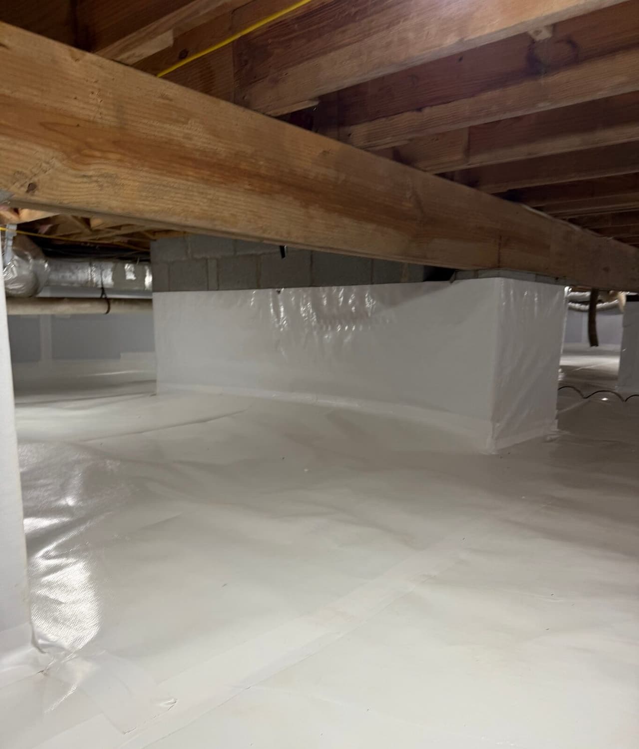 Crawl space with white liner sealed over the floor and wrapped around a block pier below a wood beam.