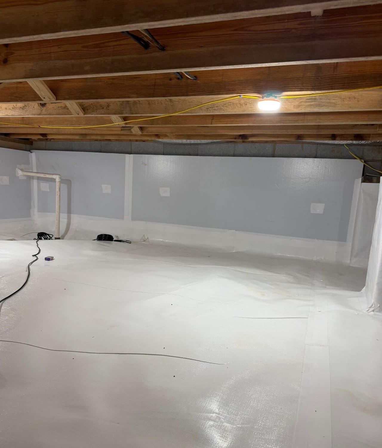 Wide view of a crawl space with white floor liner, gray foam board walls and a work light overhead.