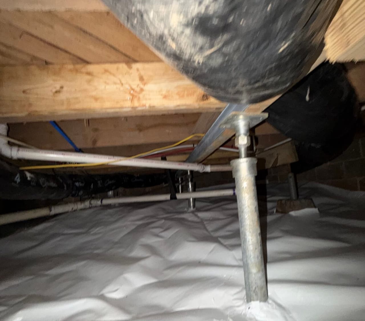 Adjustable steel support post under a joist standing on white crawl space liner, with pipes and ducts nearby.