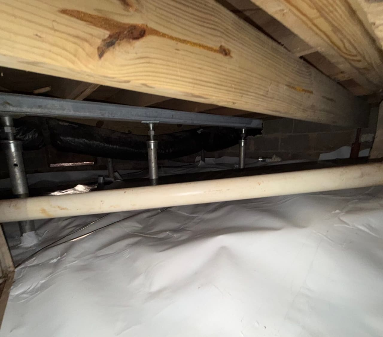 Steel beam held by adjustable steel support posts under floor joists above a white crawl space liner.