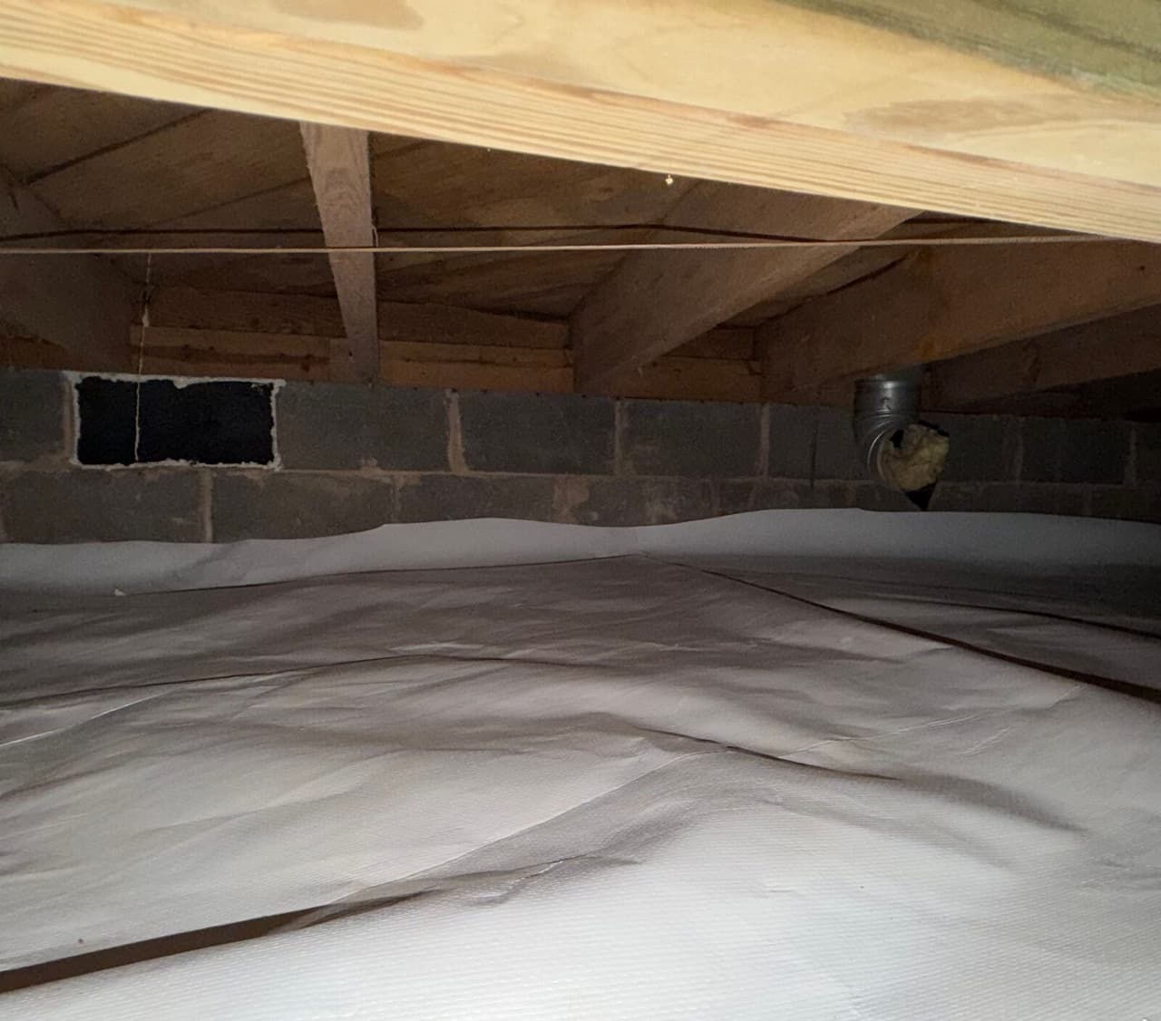 White liner covering a crawl space floor up to a block wall with a vent opening under wood joists.