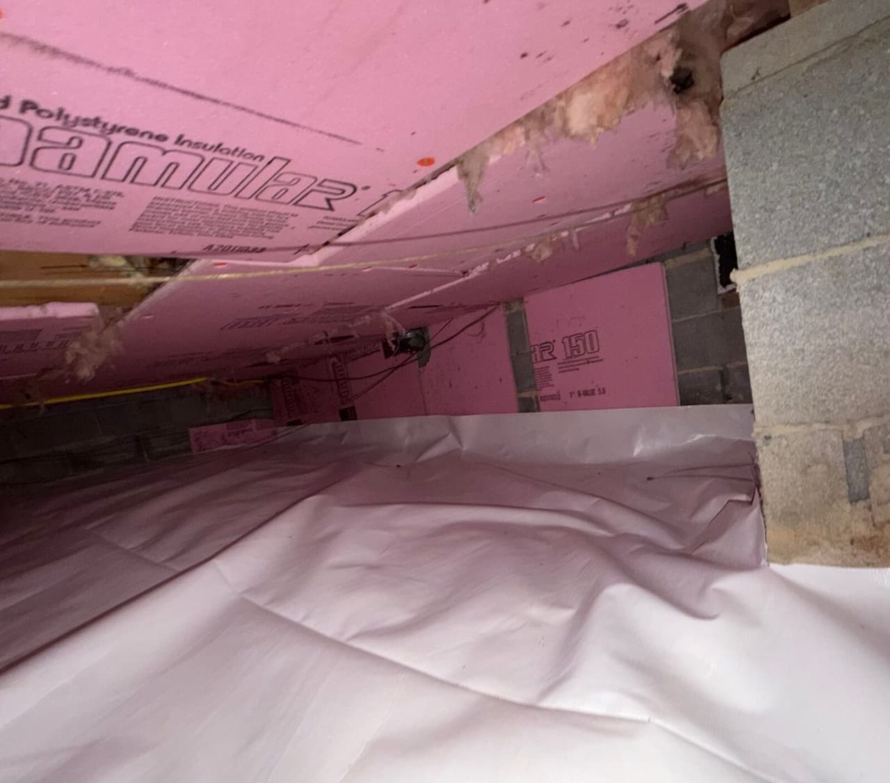White liner covering a crawl space floor beneath pink foam insulation boards next to a block pier.