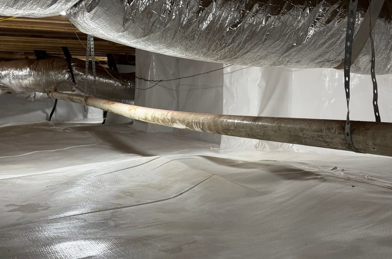 Crawl space with white liner on floor and wrapped pier under a pipe and ducts.