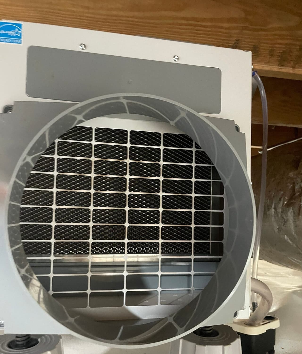 Crawl space dehumidifier mounted under floor joists with a round duct opening and a drain line attached.
