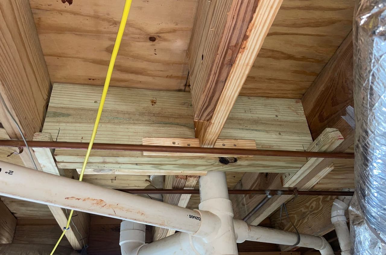 New lumber framing installed beneath the subfloor with plumbing pipes and a yellow wire running below.