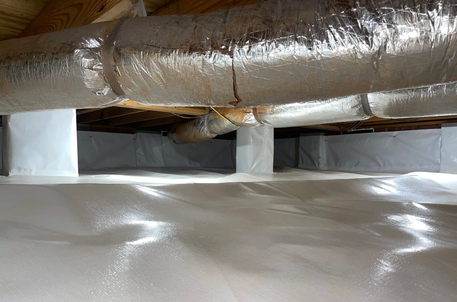 Encapsulated crawl space with white liner on the floor, walls and wrapped piers beneath insulated ducts.