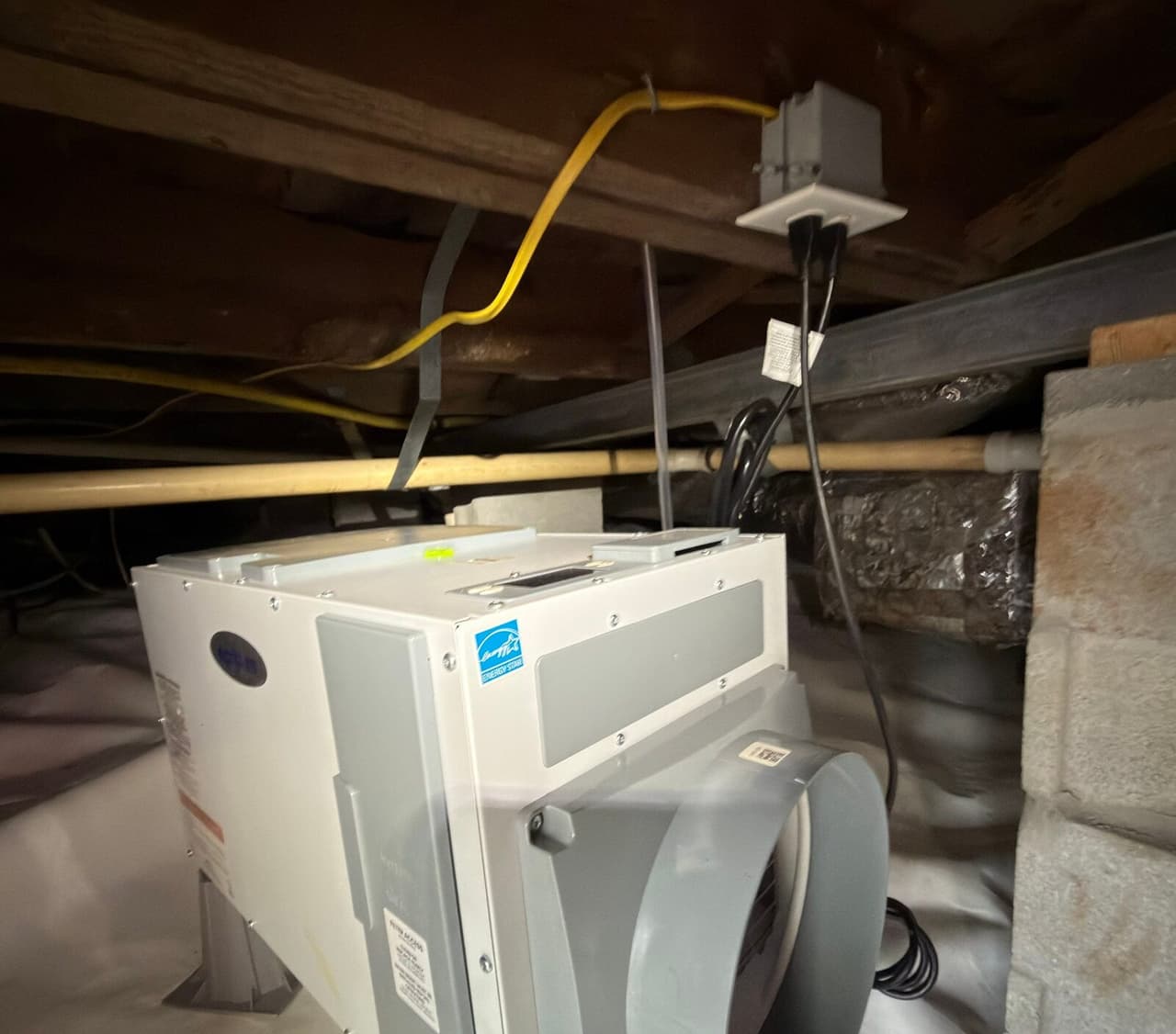 Dehumidifier on a stand in a crawl space, plugged into an outlet box on a joist, with white liner below.