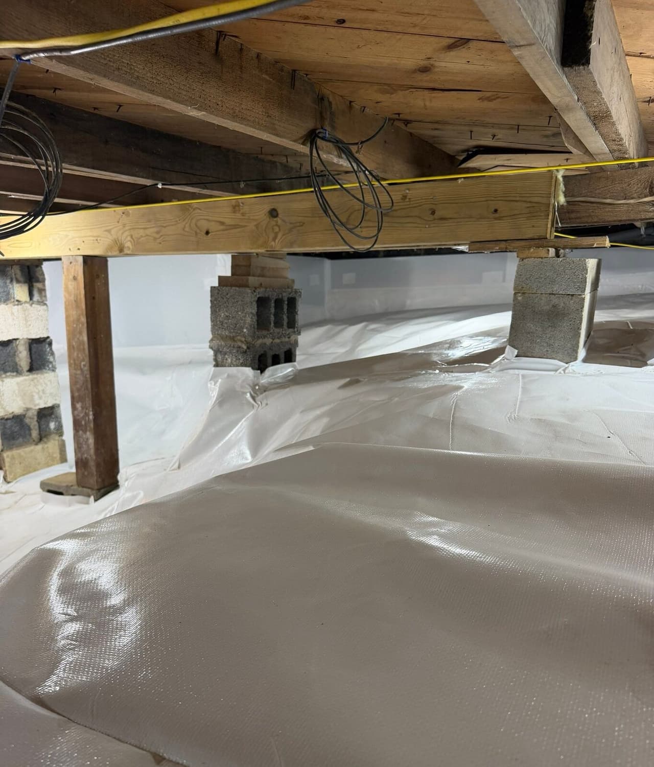 A crawl space with white liner covering the floor and wrapped around block piers under wooden beams.