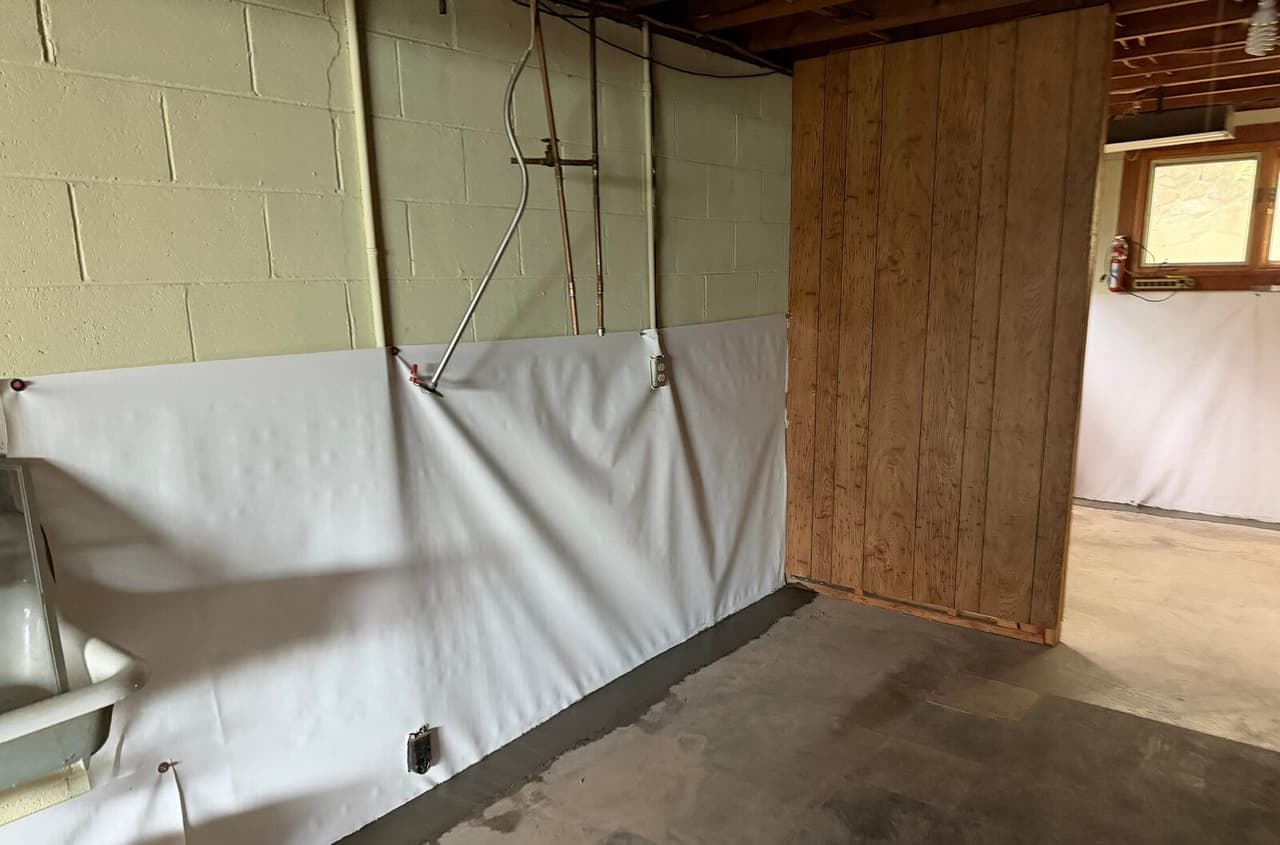 Block wall with white liner below pipes and a patched concrete strip along the floor beside a wood panel wall.