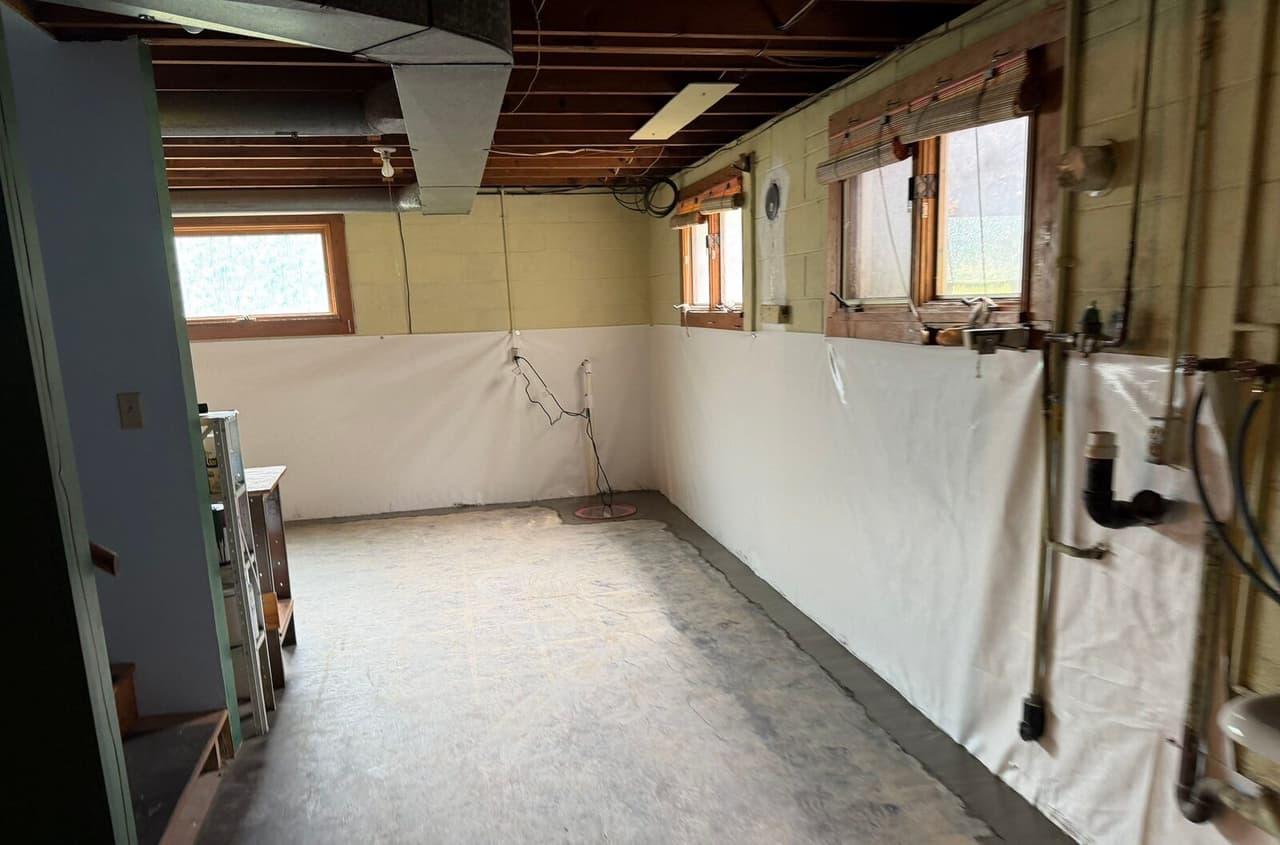 Basement with white liner on walls, patched floor edge, sump lid in the corner and pipes on the right wall.