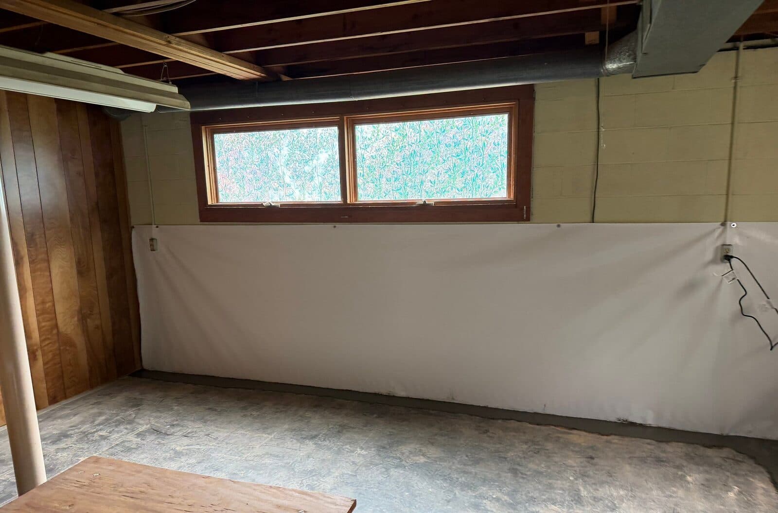 Basement wall below a window covered with white liner, with a patched concrete strip along the floor.