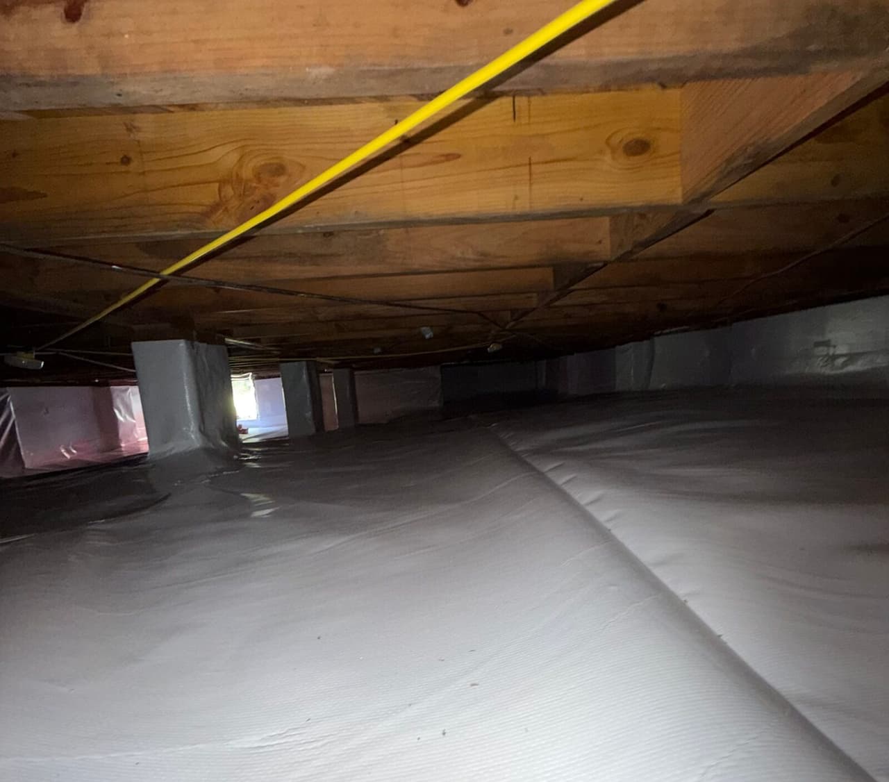 Crawl space floor covered by white liner with wrapped piers under wooden floor joists.
