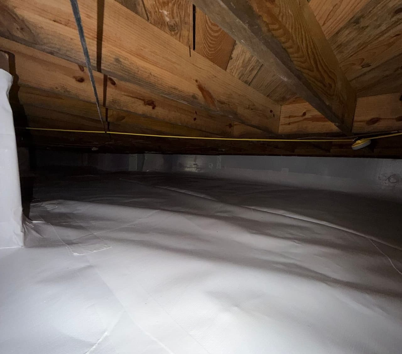 Crawl space with white liner on the floor and walls beneath wooden joists.
