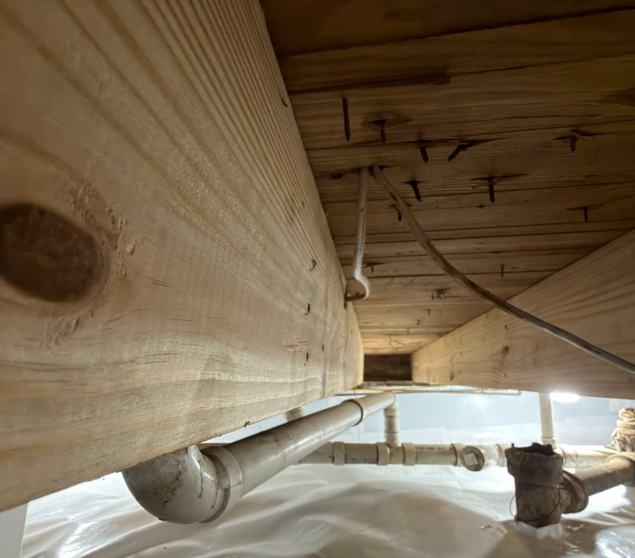 New wooden joist fastened under the subfloor above plumbing pipes and a lined crawl space floor.
